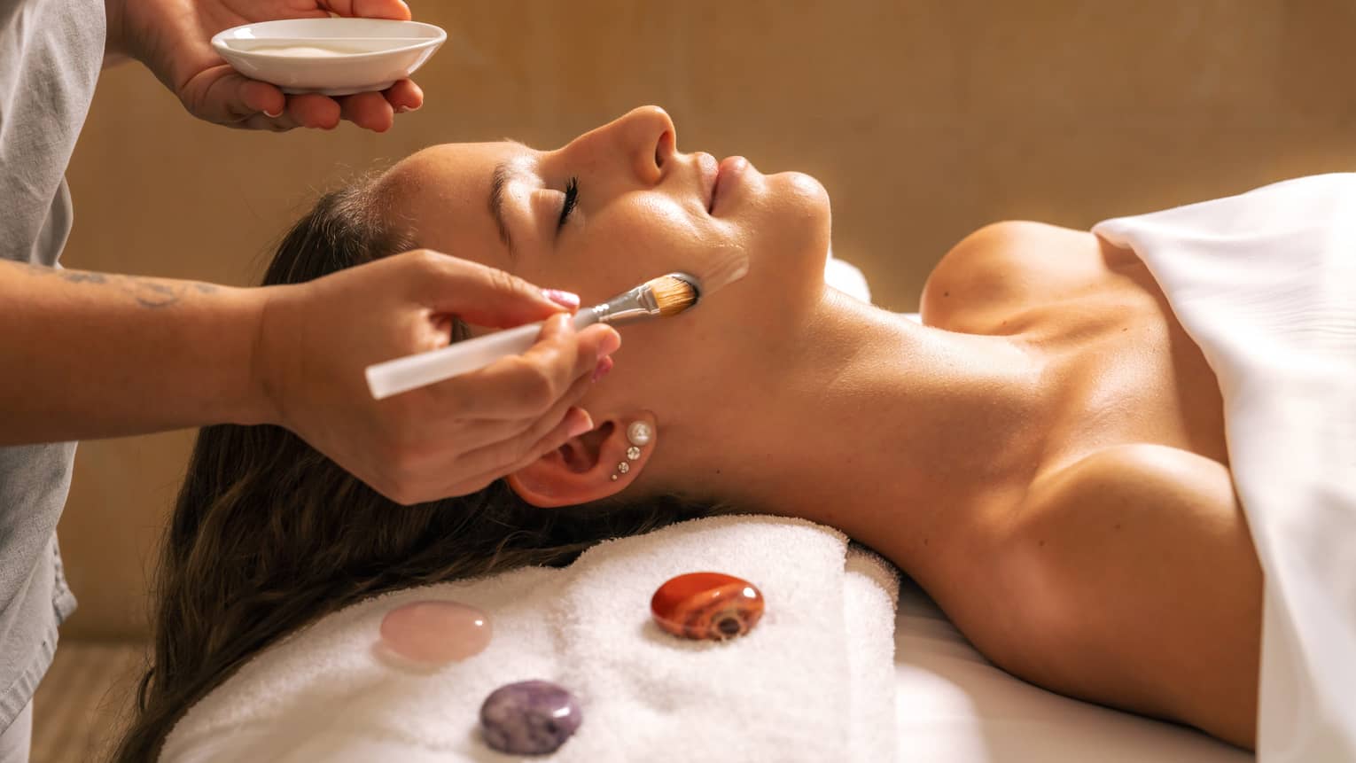 Woman lies covered by white blanket from shoulders down as someone gently applies a cream on their face with a brush, with three gemstones places next to the client's head