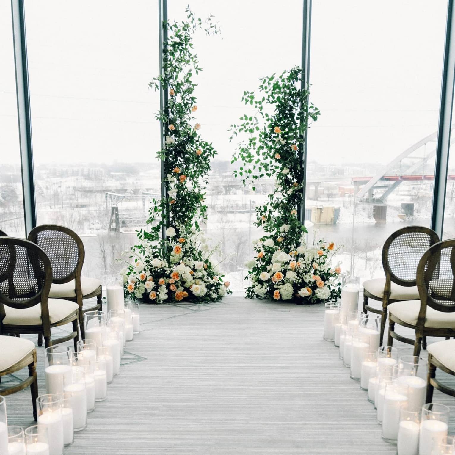 An indoor wedding ceremony set-up with tall floral arrangements, candles lining the aisle and a large floor-to-ceiling window overlooking a city skyline