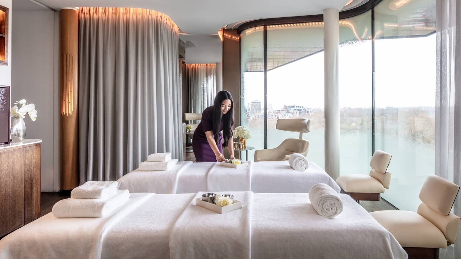 Spa couples treatment room with staff member, two white beds and chairs at ends, tall windows with city views