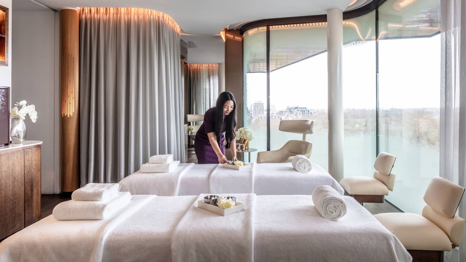 Spa couples treatment room with staff member, two white beds and chairs at ends, tall windows with city views