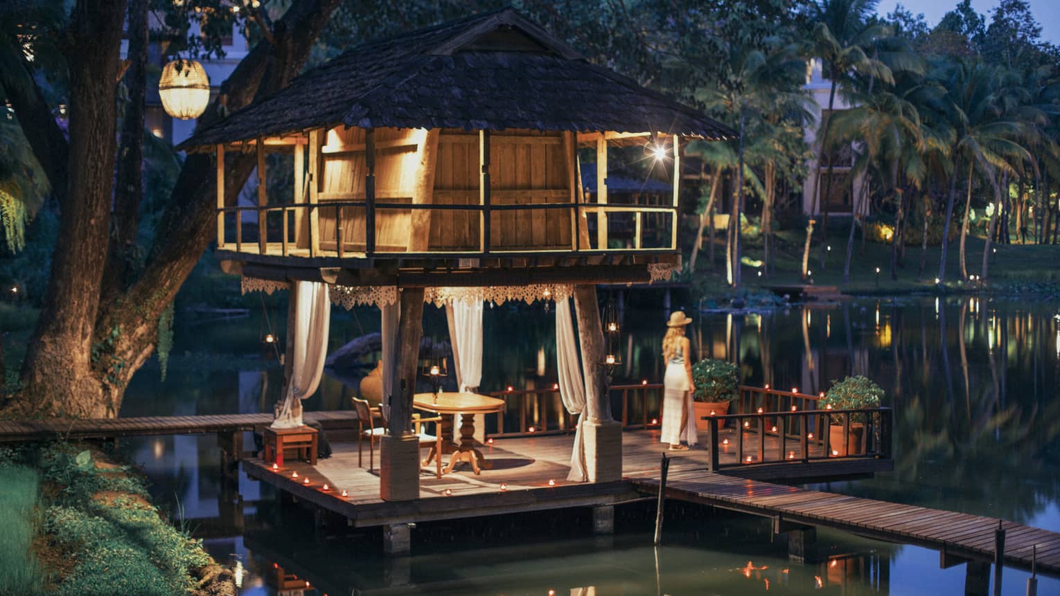 A guest looks out at a serene river at dusk, on the platform of a wooden hut, as lanterns and twinkling lights dot the scene.