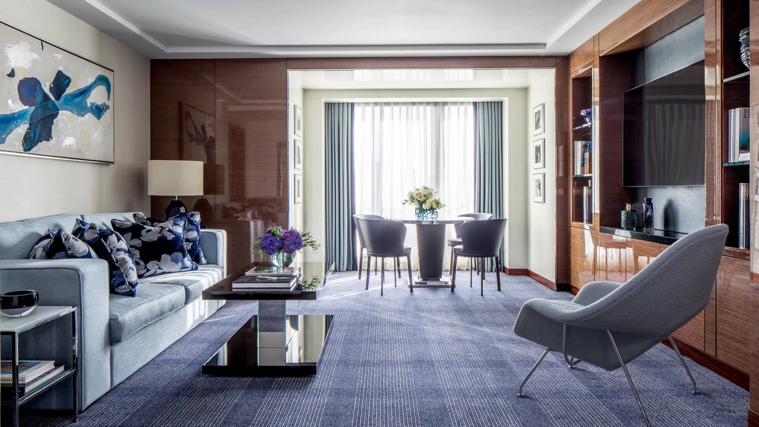 The Ambassador Suite at Four Seasons London Park Lane in Mayfair features a sophisticated and spacious living area. The room is elegantly appointed with rich wooden paneling and a deep blue carpet that contrasts beautifully with the light grey modern sofa and chairs. A large abstract blue butterfly painting dominates one wall, adding a vibrant splash of color. The space includes a sleek, dark coffee table and a dining area in the background with a round table and chairs, positioned near a bright window draped with sheer curtains.