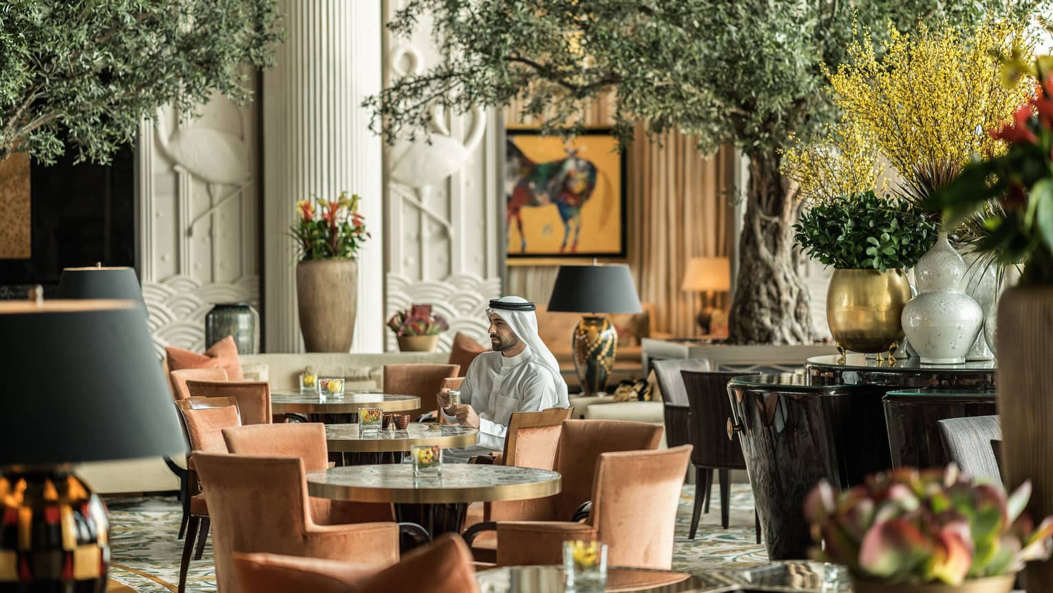 Man in traditional Bahraini dress sitting in lounge, surrounded by round tables with rust chairs and lush greenery