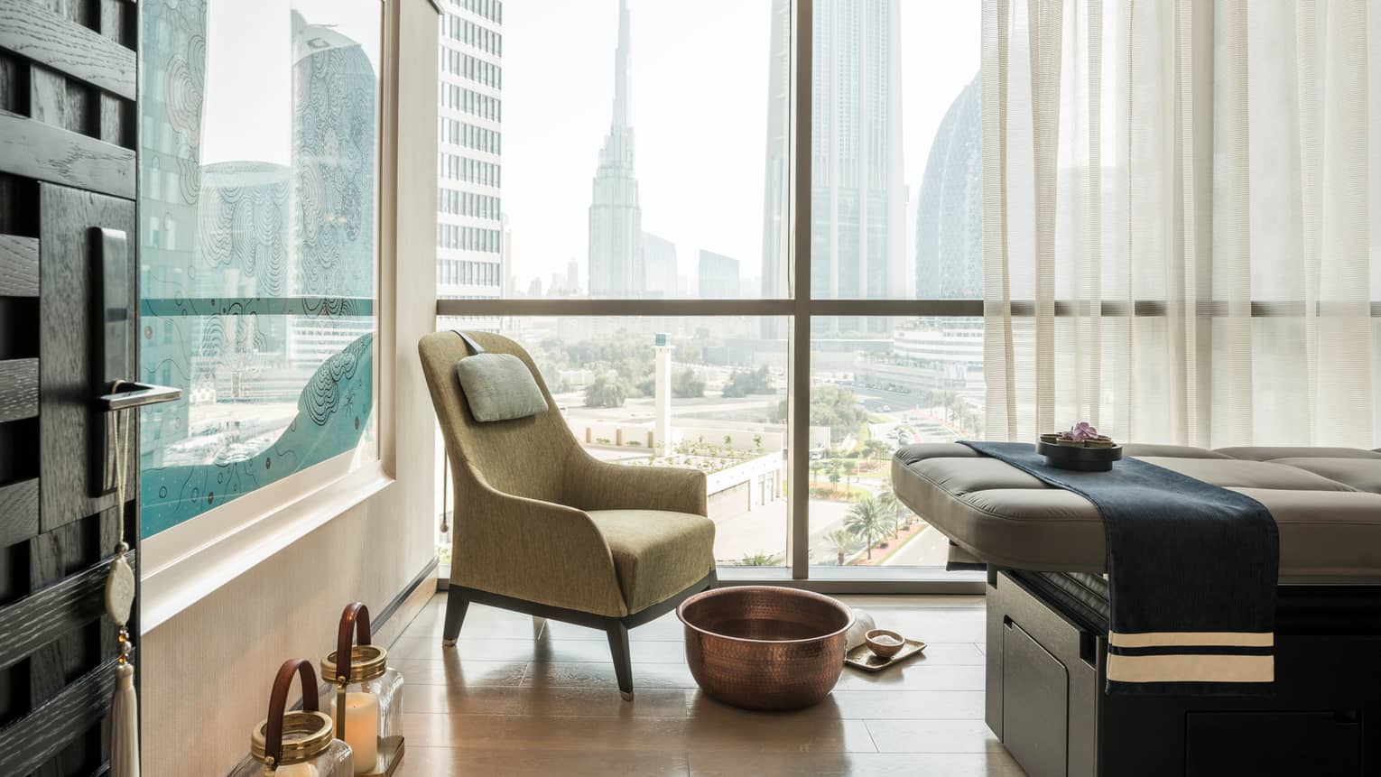 Armchair in hotel room corner by sunny window, large brass bowl foot bath, rolled white towel on floor