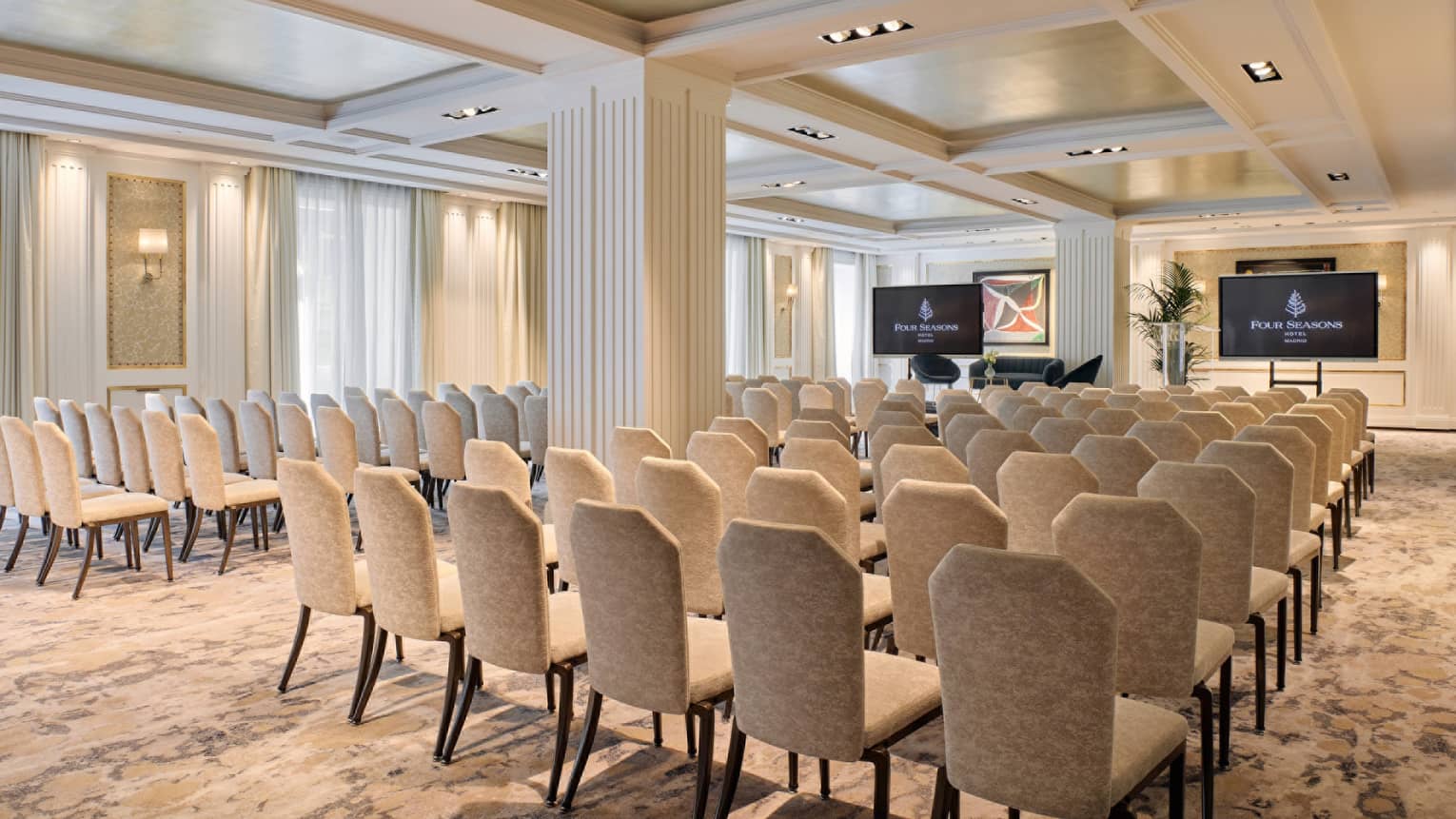 Light-filled meeting room with large windows, beige walls and carpet, and rows of chairs facing two flat-screen TVs on the wall