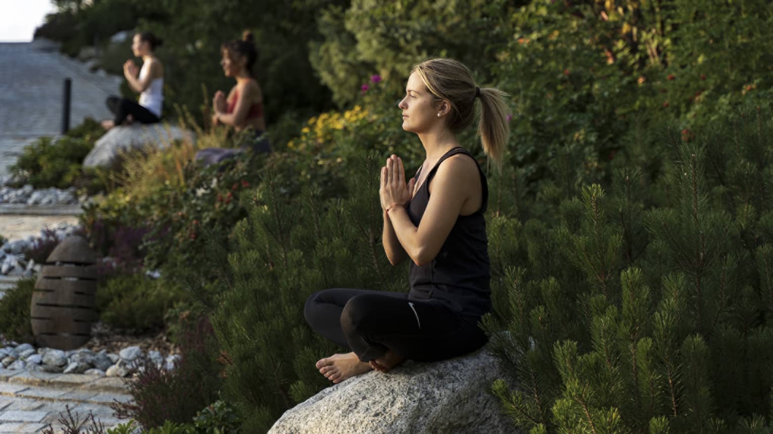 Three women outside in seated yoga pose on boulders separated by flowers