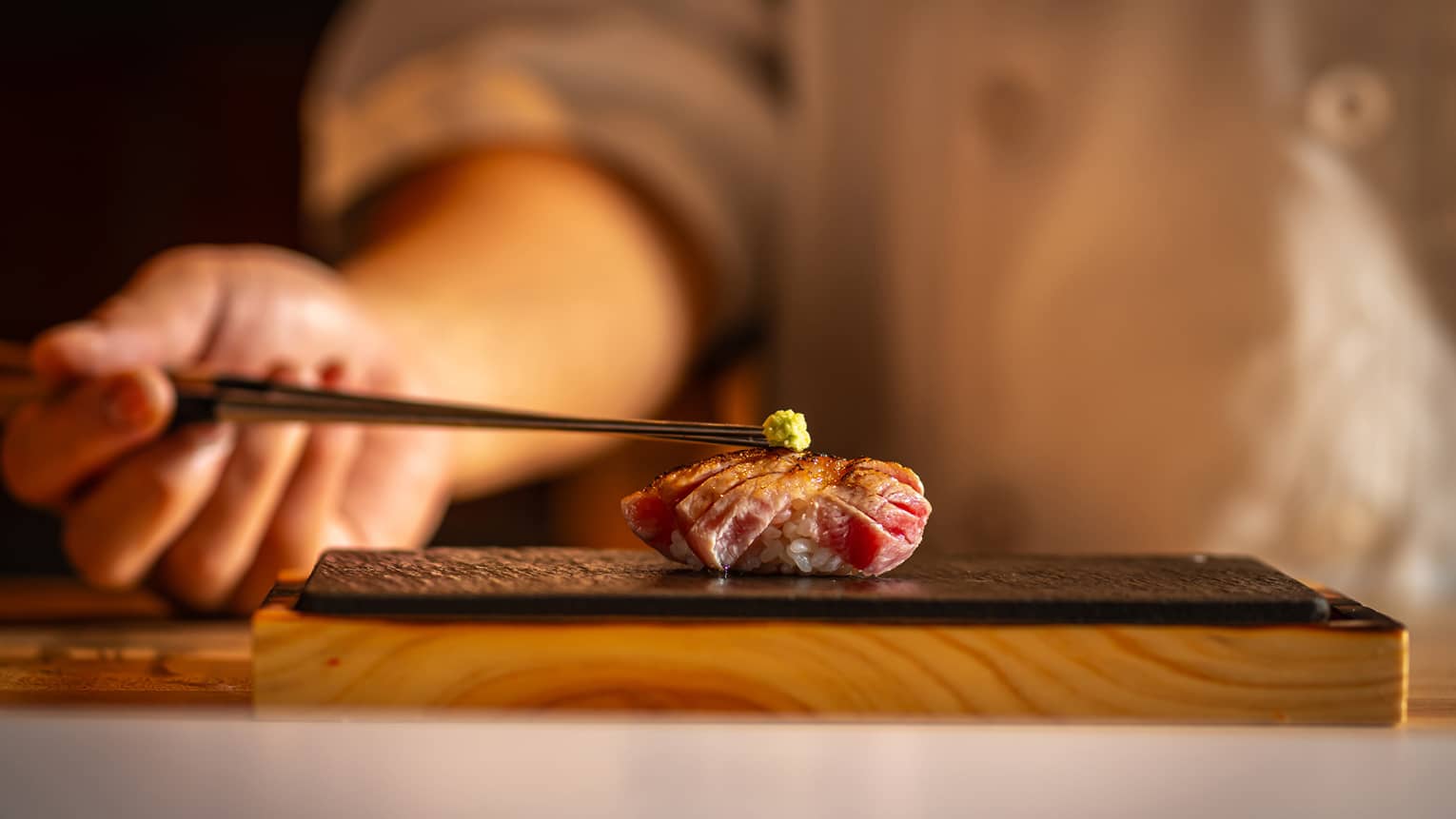 Seared tuna nigiri topped with a small dab of wasabi, placed on a black slate plate with a warm, blurred background