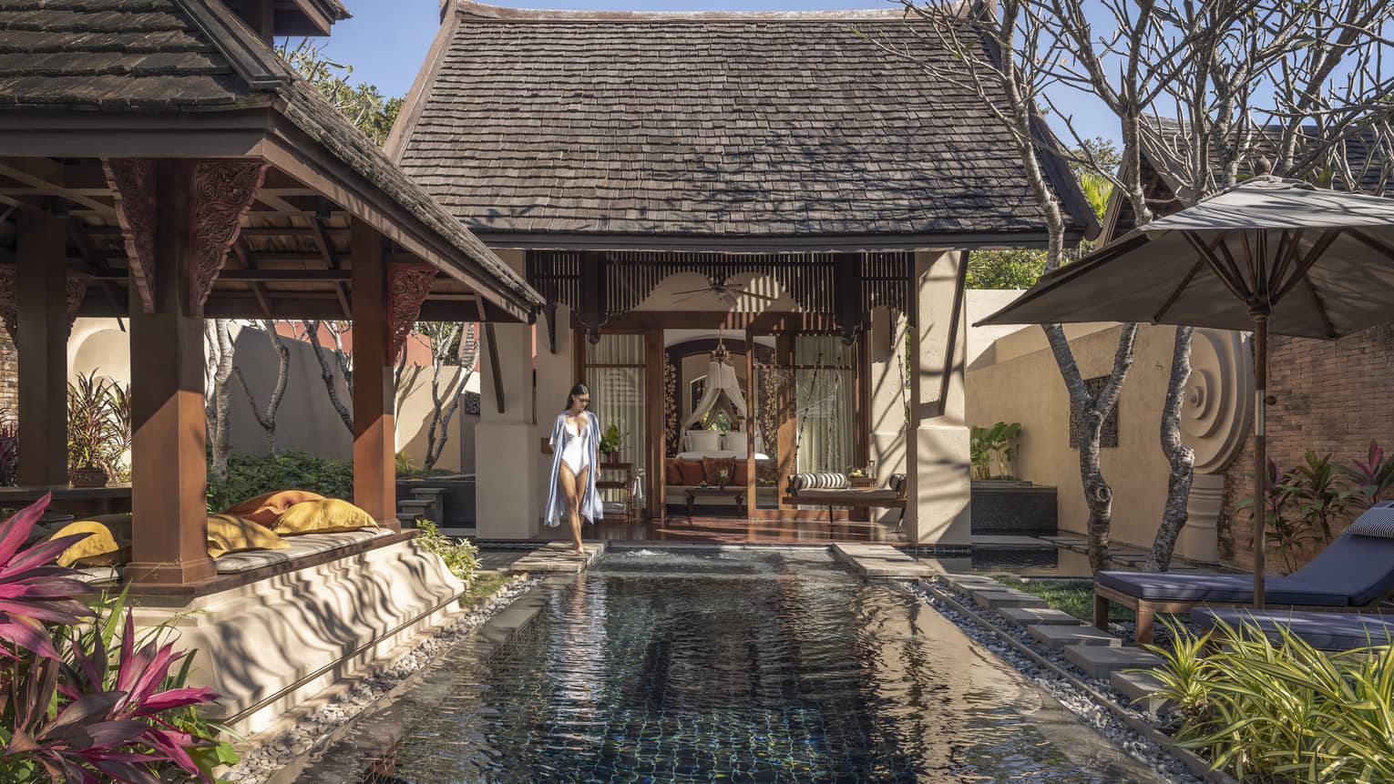 Person walking beside a private pool in a courtyard with sun loungers, tropical plants and traditional wooden architecture