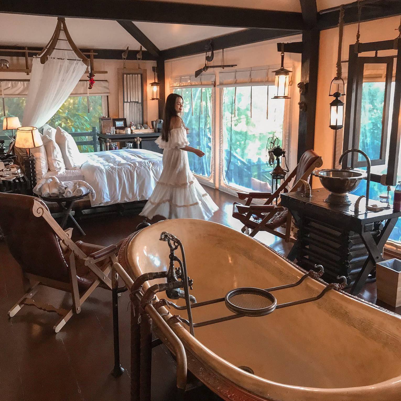A woman wearing a white dress stands by the windows of a hotel room