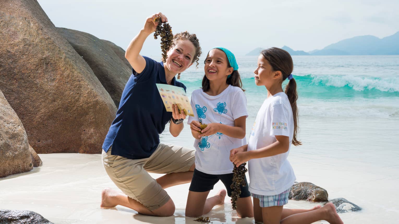 Woman kneels on white sand beach beside boulder, holds up seaweed to show two girls