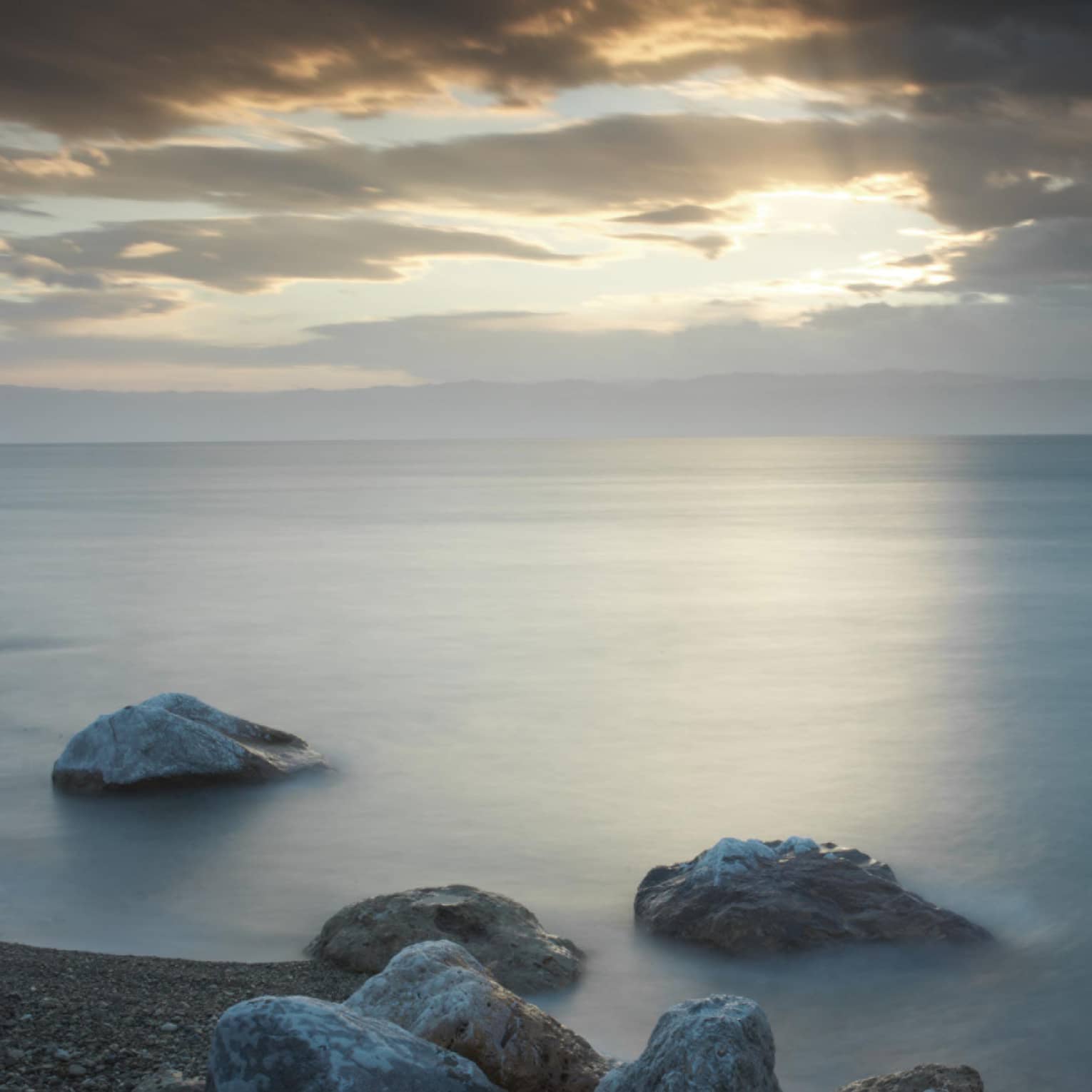 By a stony shoreline, rocks jut out of a dark lake beneath a beam of sunlight filtering through a hole in dark grey clouds.