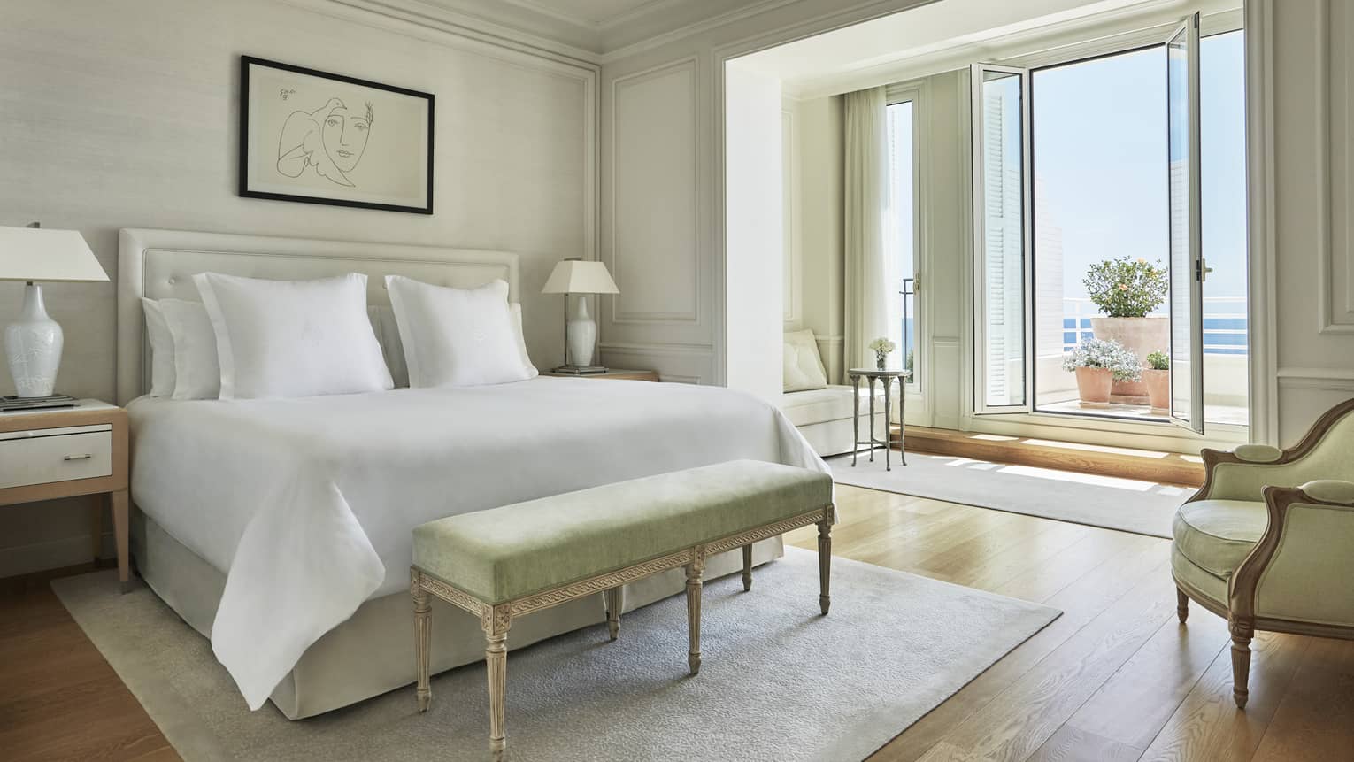 Hotel suite with white bed and green foot bench on neutral rug with terrace and sea views from tall glass terrace doors