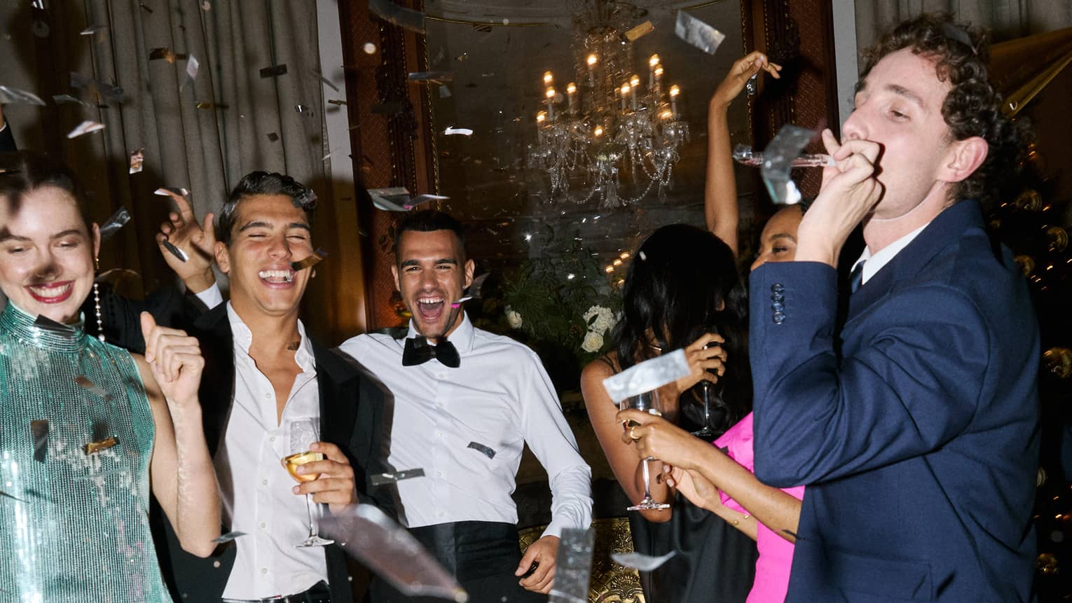 Six people dressed in cocktail party attire smile and celebrate as metallic confetti falls over them in a hotel suite