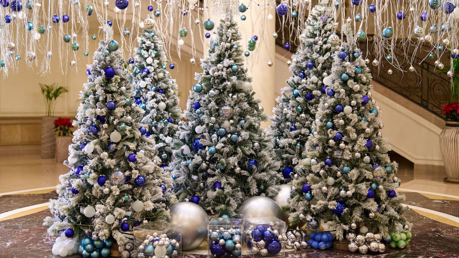 A lobby is decorated with hanging white garland and five frosted Christmas trees adorned with blue and silver ornaments.