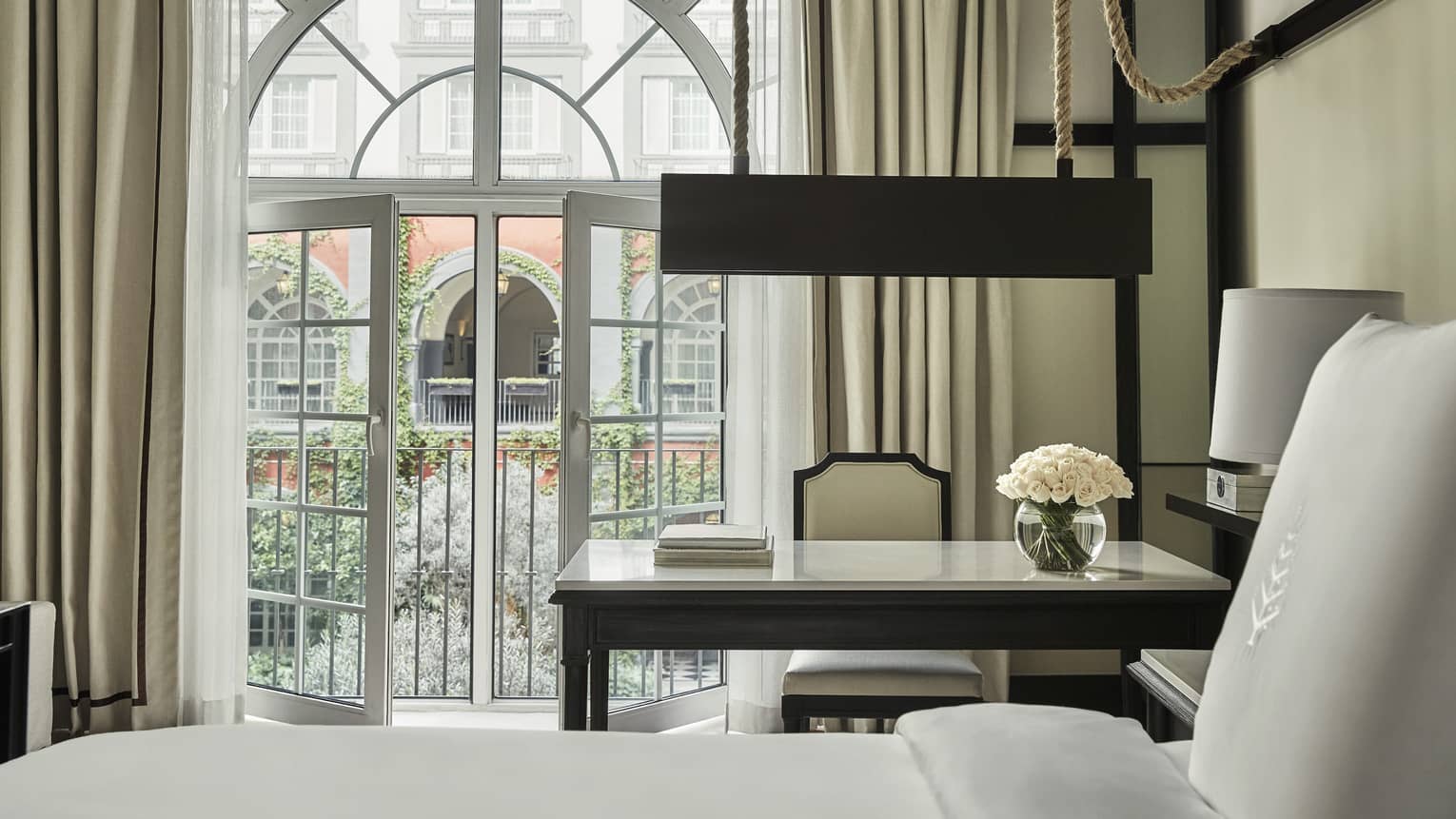 Elegant hotel room with a large arched window, a desk with flowers, and an inviting view of a garden courtyard framed by ivy-covered walls.