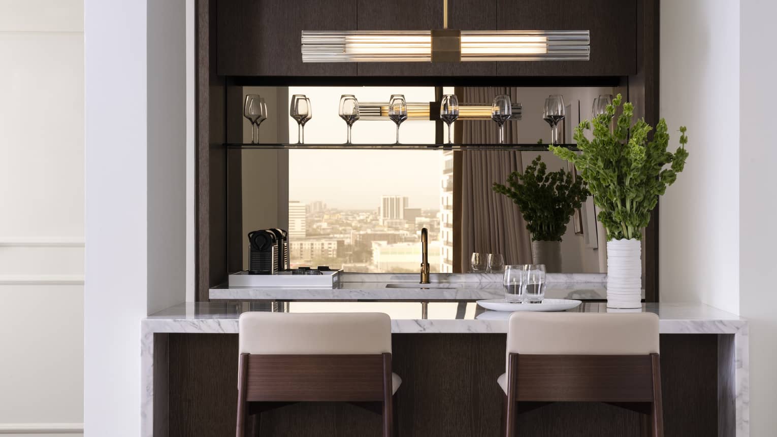 A kitchen with wine glasses on a shelf, herbs, two bar stools, an island and sink.