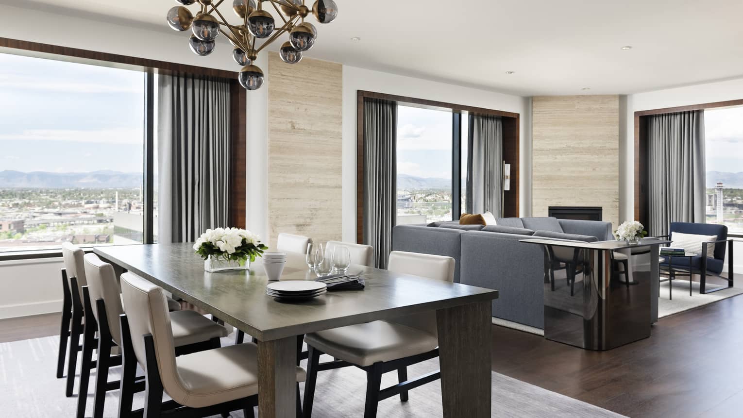 A large rectangular dining room table with a gold chandelier next to large windows and a seating area with sofa and chairs.