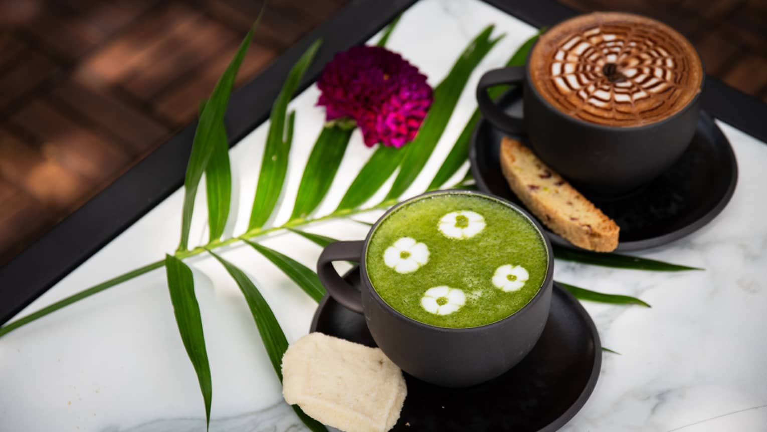A black mug of coffee with a latte and brown-and-white foam and another black mug with tea with green foam and four white floral patterns