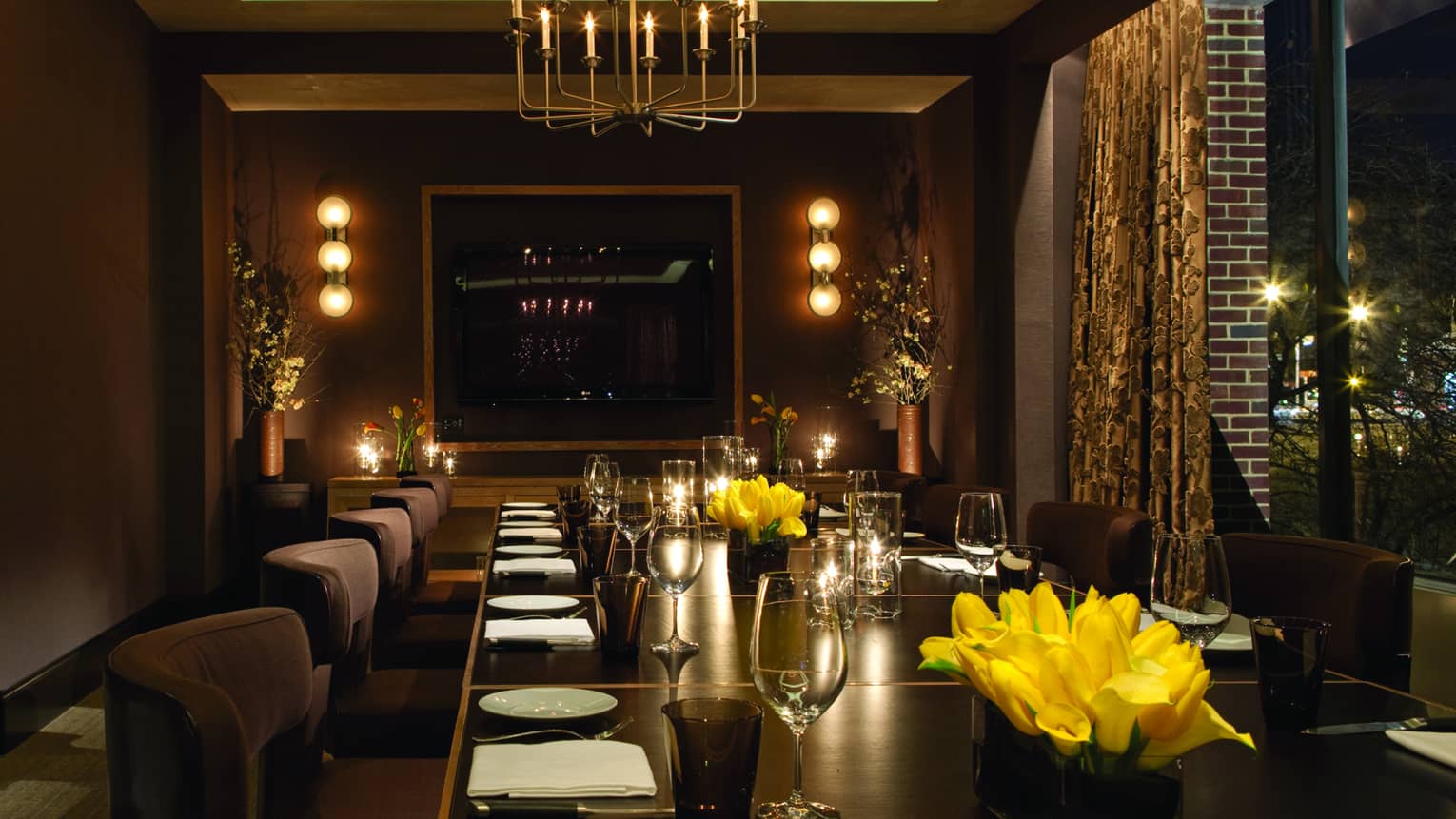 Long private dining table with plush chairs set with wine glasses, yellow flowers, candles, low lights on walls