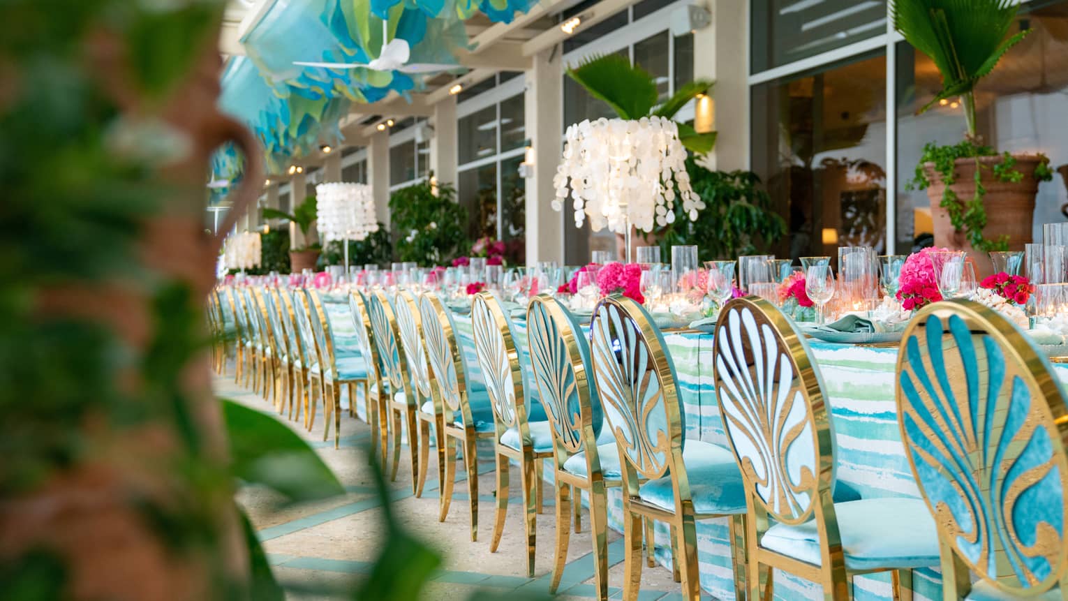 A row of turquoise & gold chairs at a long table full of pink floral arrangements, white chandelier, palm tree accents 