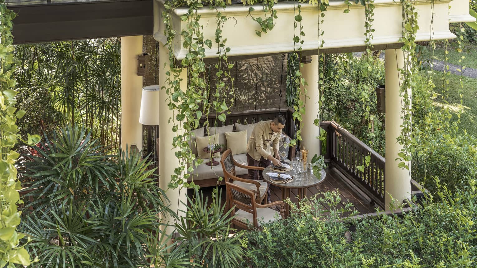 Person setting a dining table in a covered outdoor seating area surrounded by hanging plants and lush greenery