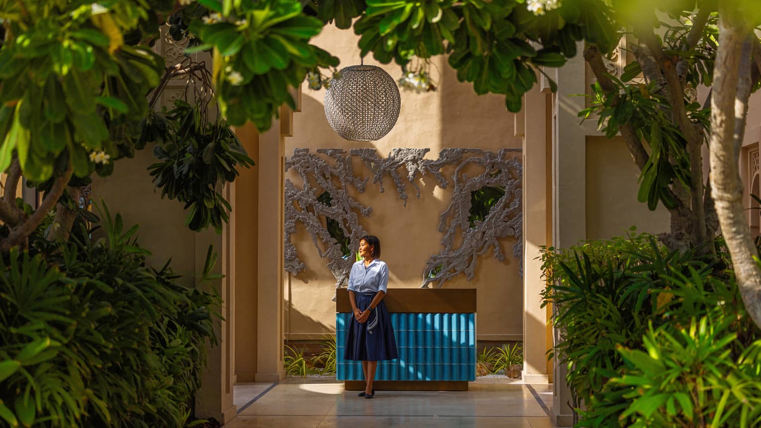 Woman staff member wearing light-blue button down shirt and dark blue knee-length skirt stande next to a blue-tiled counter set surrounded by bright green plants and trees