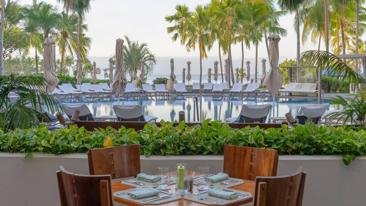Outdoor restaurant square table with four wooden chairs, set next to a tropical resort pool with ocean view