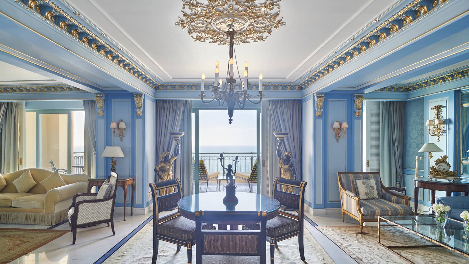 Presidential Suite dining/living area with round dining table, chaise longue, cream couch, blue wall accents, doorway to veranda with sea views