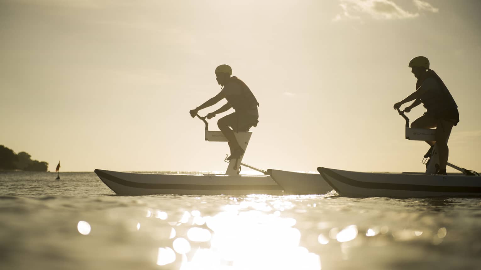 Silhouettes of two men riding aquatic bicycles on ocean at sunset