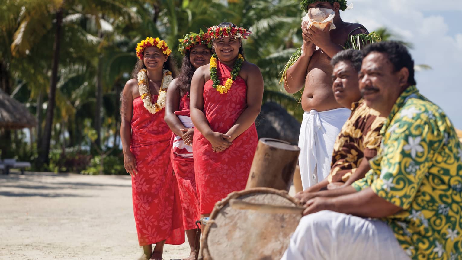 Group of people in traditional attire with leis and a conch shell, standing on a beach