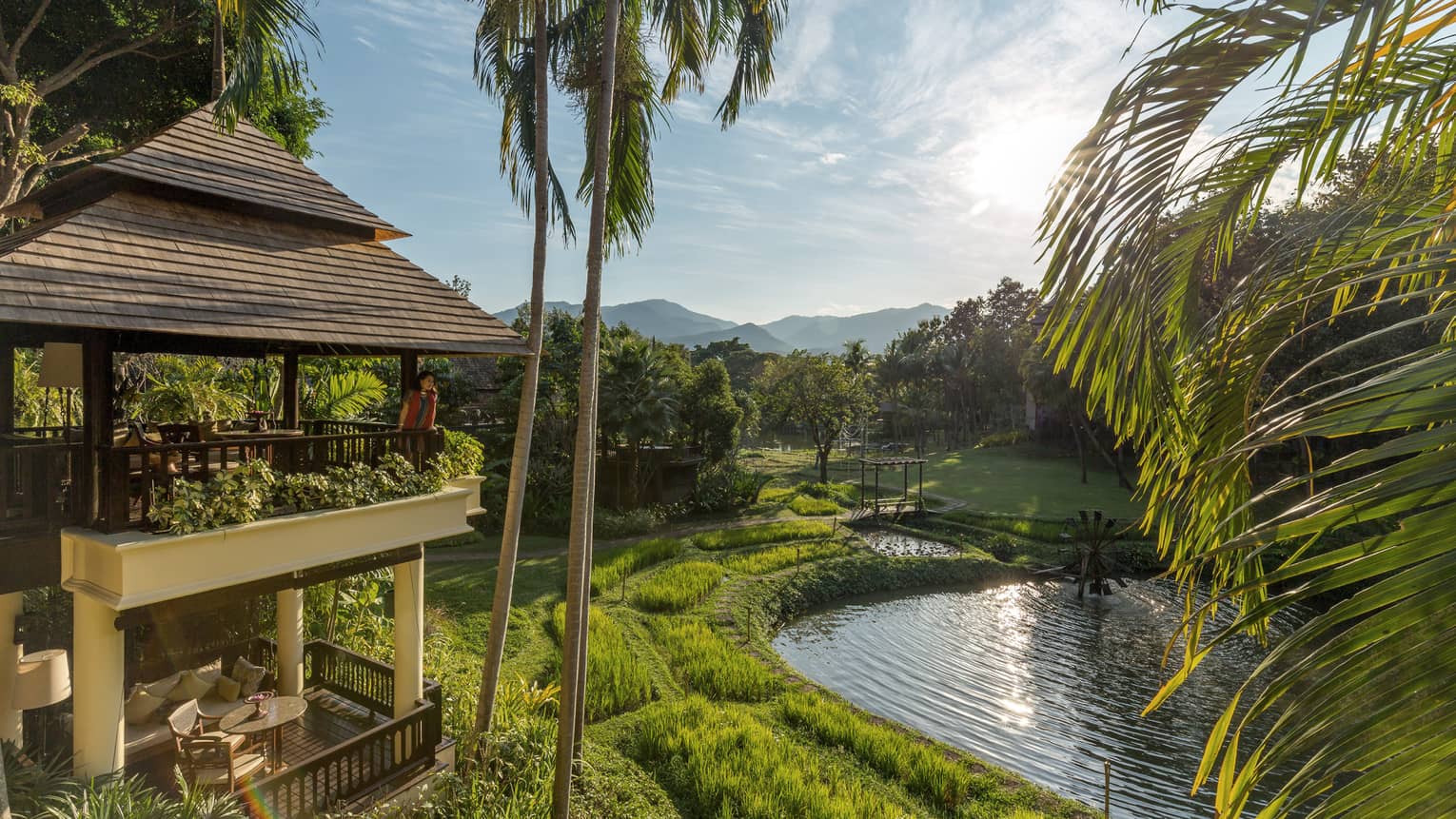 Woman stands on second floor balcony, looks beyond palms to green grass, river below