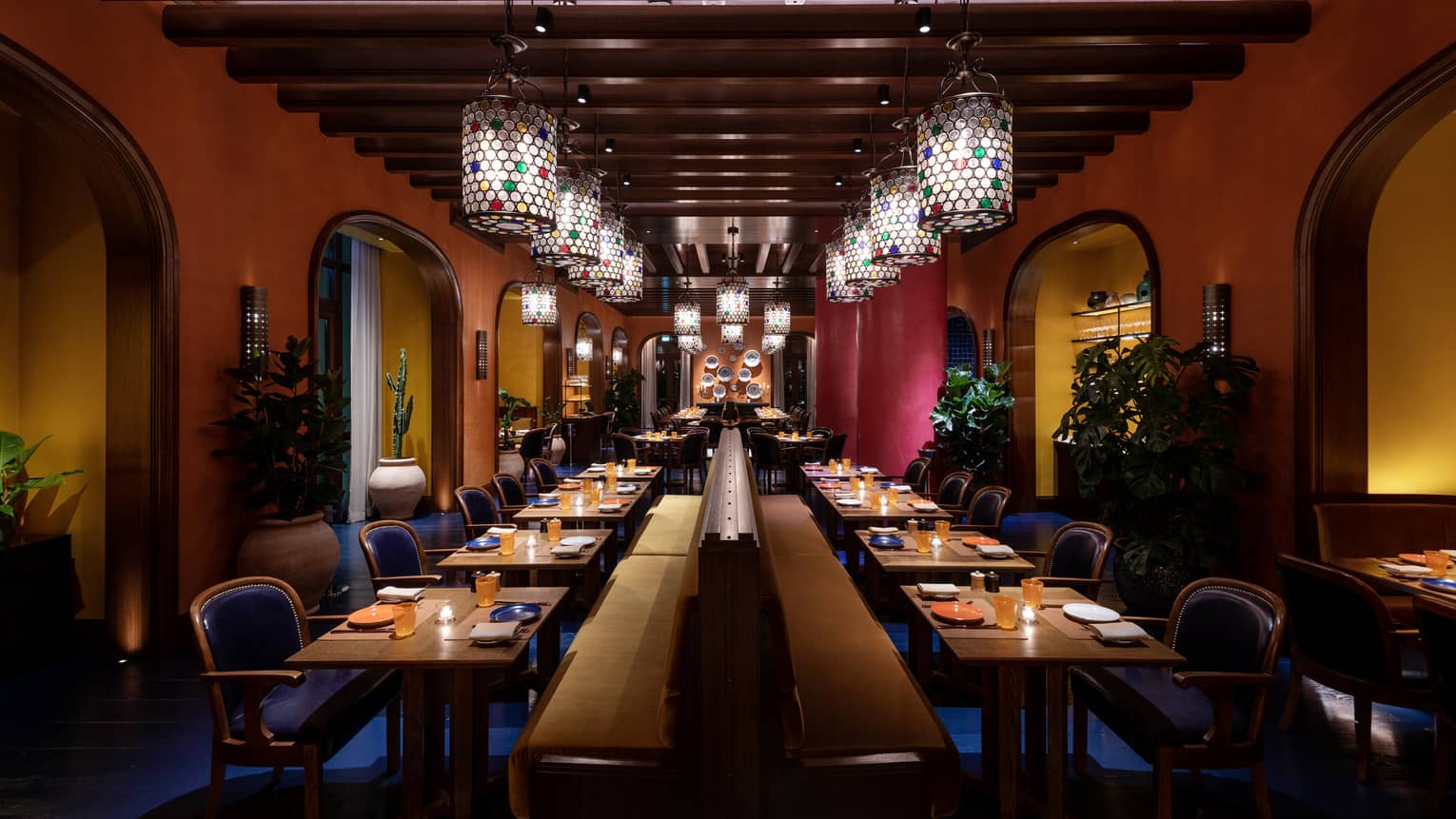 A dimly lit dining room with wood-beam ceilings and cylindrical glass chandeliers is filled with two rows of tables lining a centered banquette with rows of blue chairs on either side