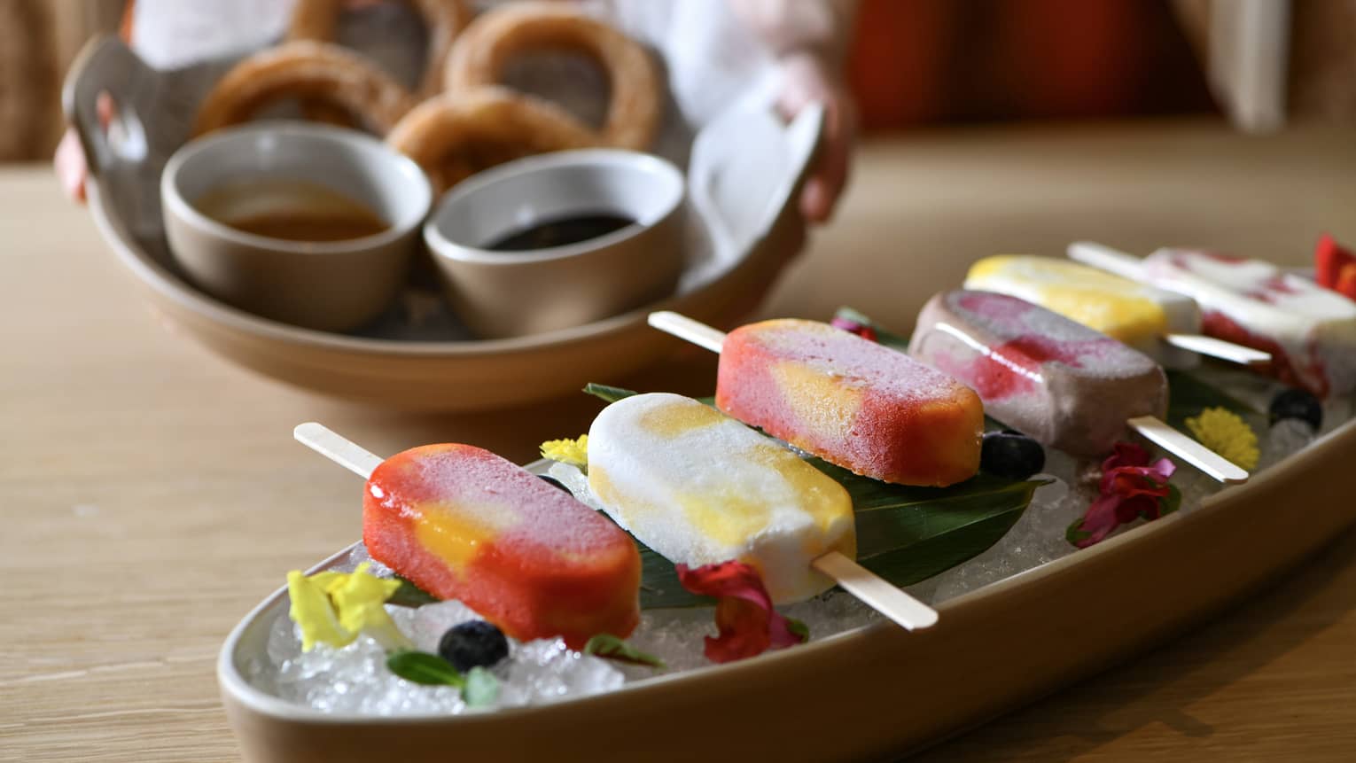 Six popsicles in alternating colors of red and white are lined up on a platter of ice and garnished with green leaves and red and yellow flower petals