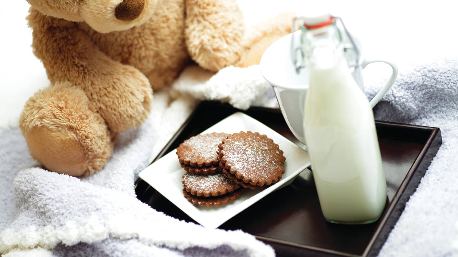 A teddy bear next to a tray of milk and cookies.