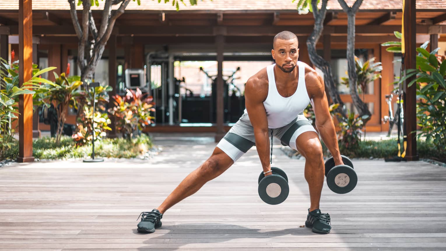 Man in white tank top and shorts doing side lunge on outdoor decking, holding weights