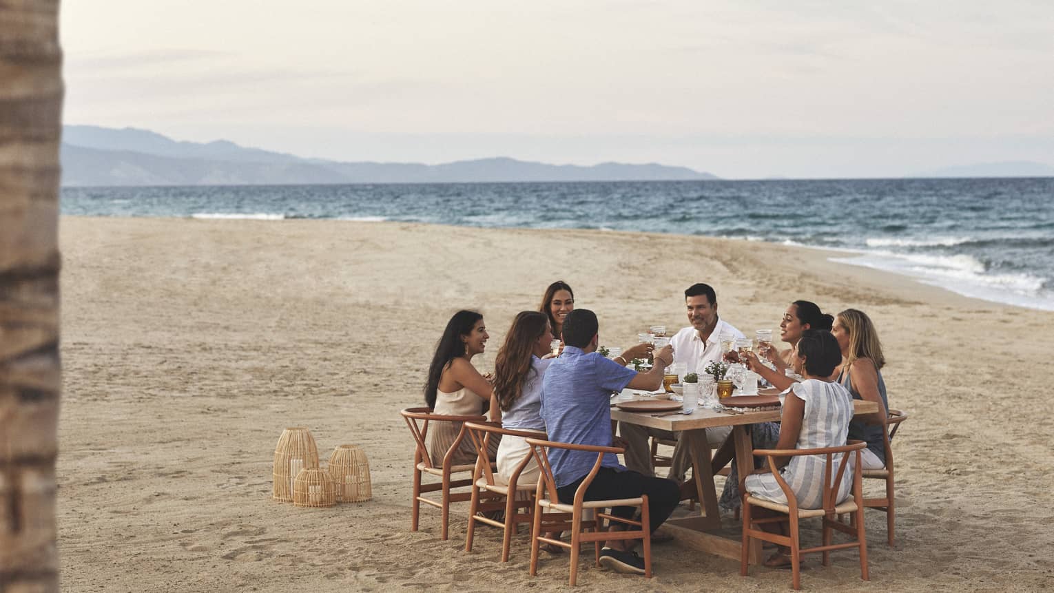 A group dines on the beach in wooden mid century modern dining chairs pulled up around a rectangular wooden dining table