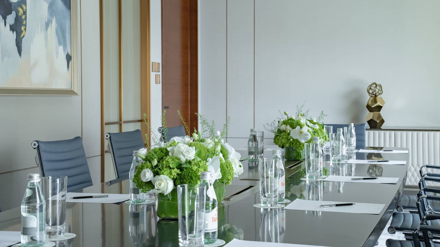 Meeting room with long boardroom table set with papers and pens, floral arrangements, bottles of water