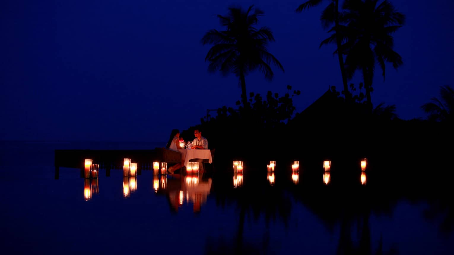Couple dine by lagoon at small table surrounded by glowing candles at night