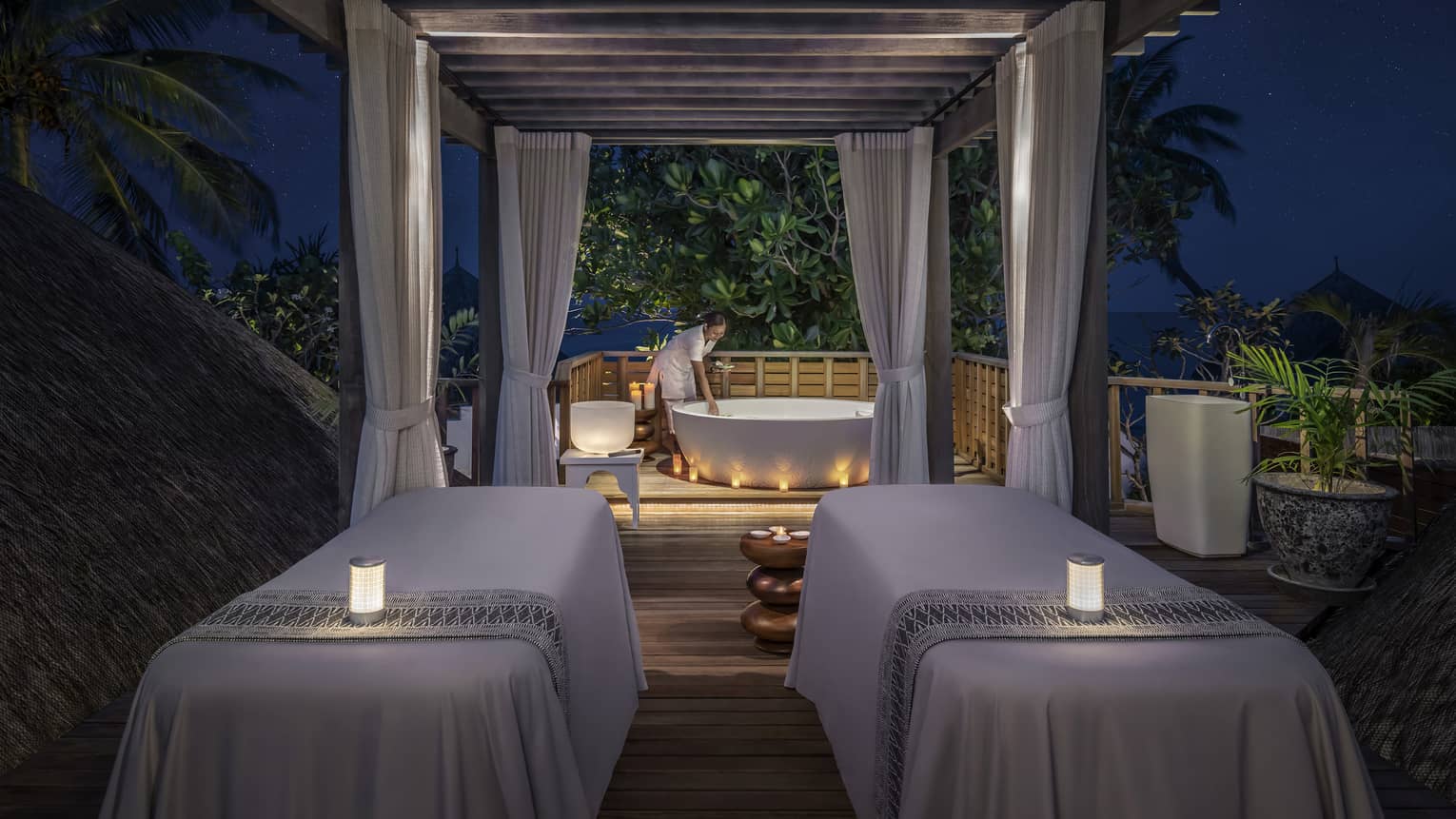 Outdoor night spa setting with two massage tables, soft candlelight, and a staff member preparing a bathtub under a canopy surrounded by tropical greenery