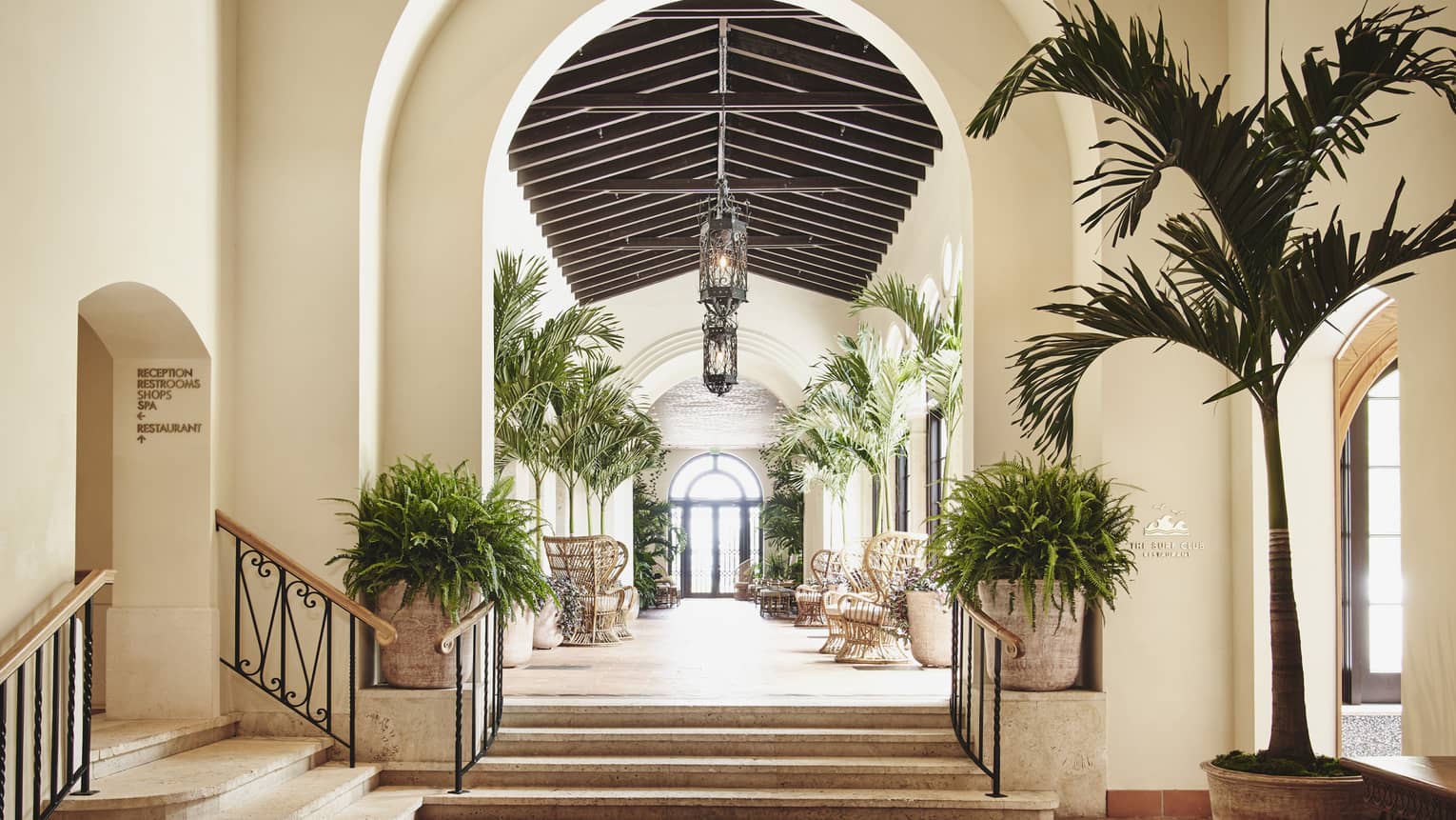 A white archway leading into a hallway with plants on both sides and large lamps hanging from the ceiling.