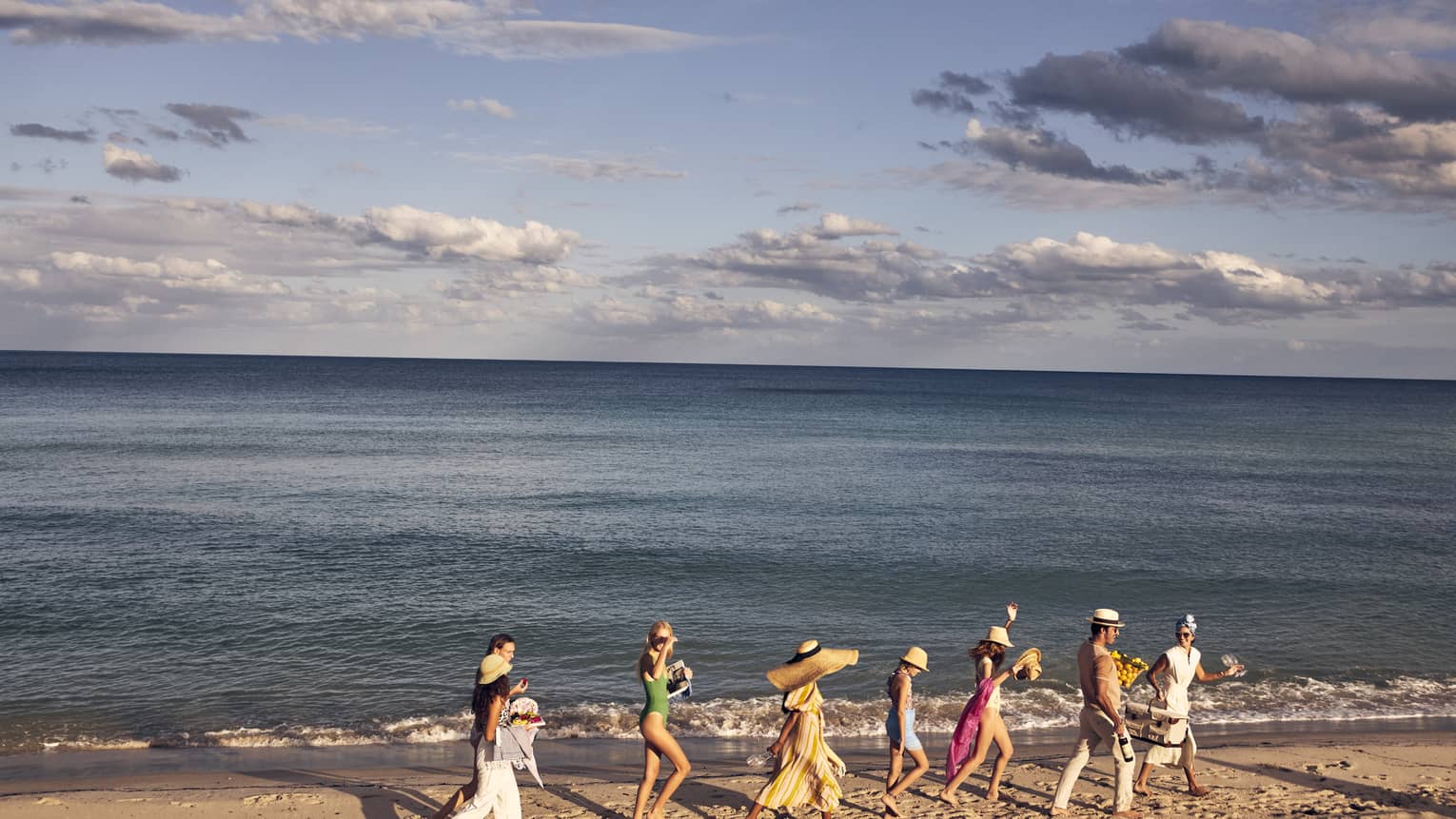 A group of people walking along a sandy beach by the ocean, wearing summer clothing and hats, with a backdrop of a calm sea and partly cloudy sky.