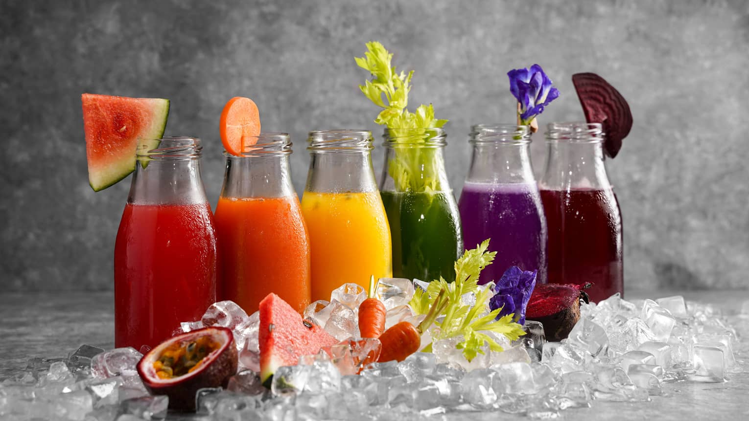 Six bottles of assorted colourful juices and garnishes lined up behind a pile of ice cubes