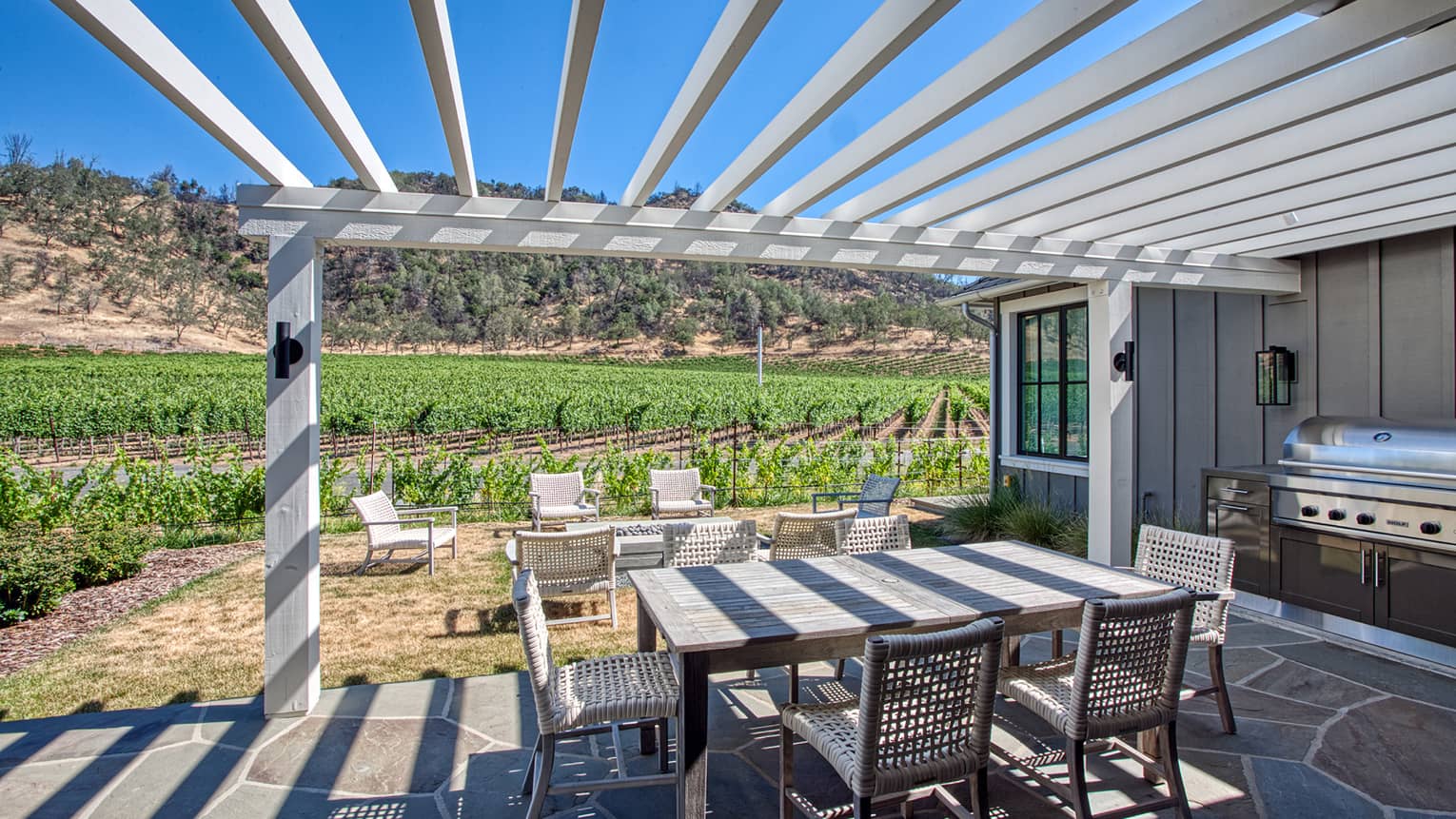 Stone terrace with dining table and grill under a pergola looking out to vineyards