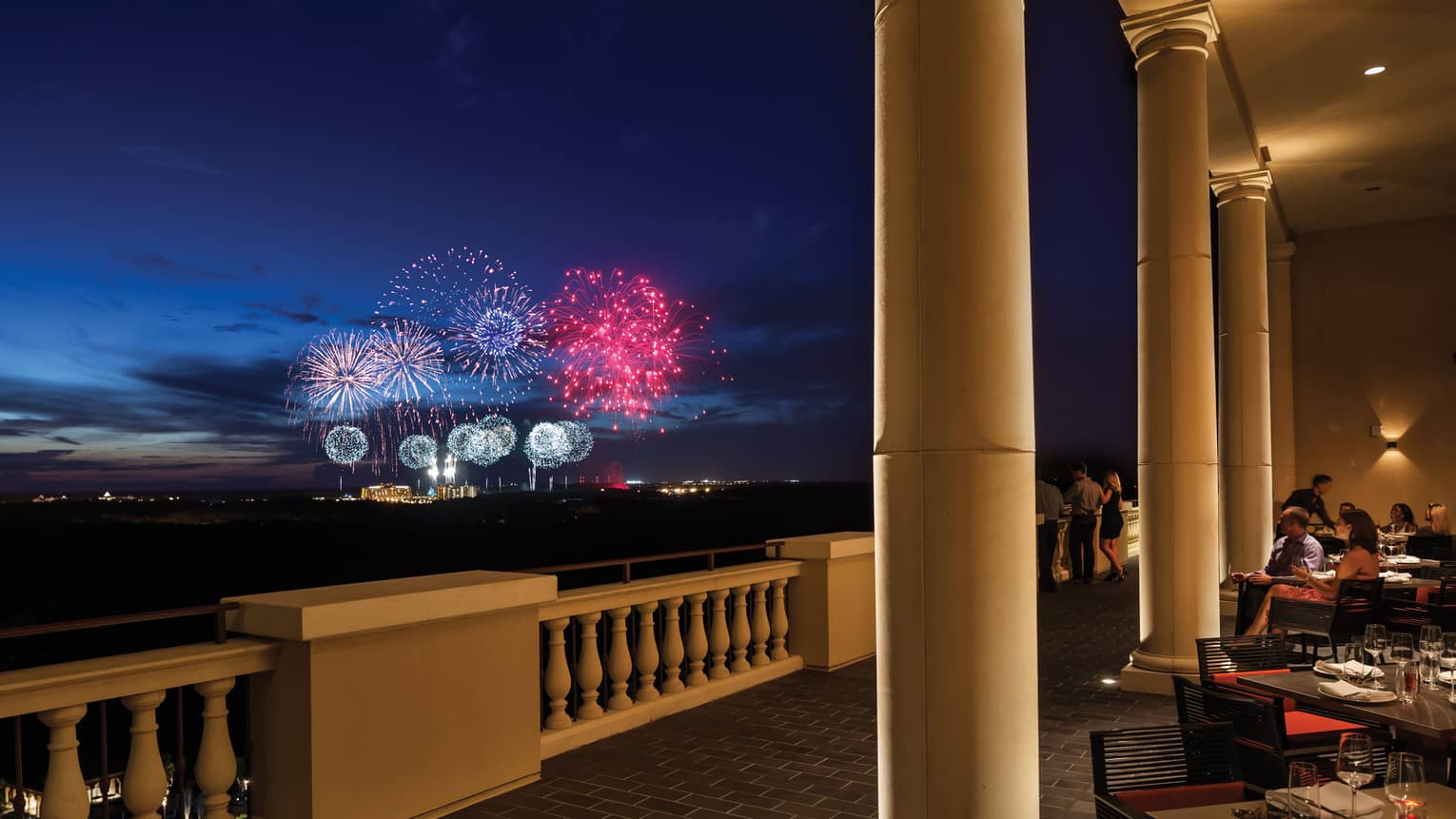 The terrace of a restaurant with tables and people watching fireworks in the distance.