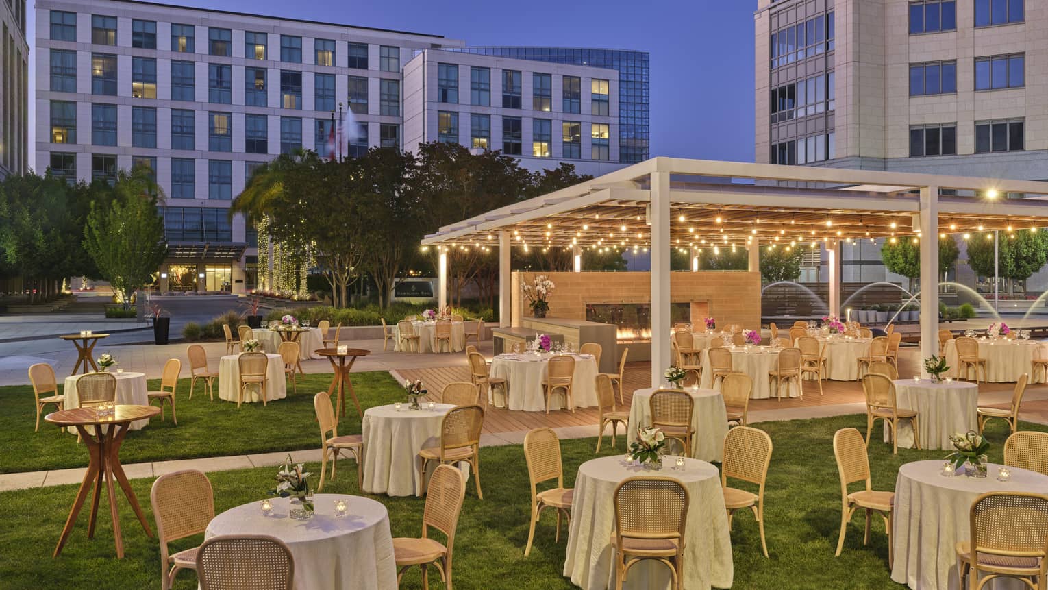 Courtyard with event setup of small round tables, some under a lit pergola, with hotel in backdrop at dusk
