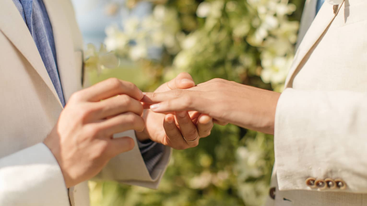 A closeup of a man putting a wedding ring on another man's hand, there are green plants in the background.