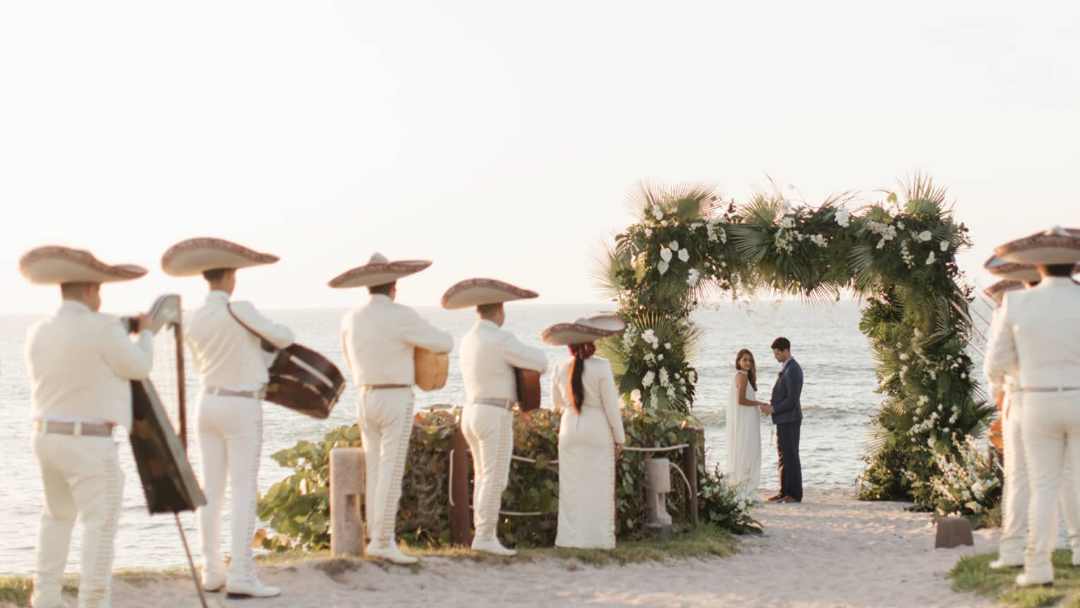 A bride and groom are standing underneath an archway of flowers and plants while men and women in white suits and large hats play music in front of them.