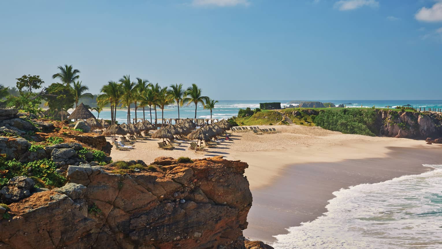 White sand beach lined with lounge chairs, palms, turquoise tides from oceanside cliff