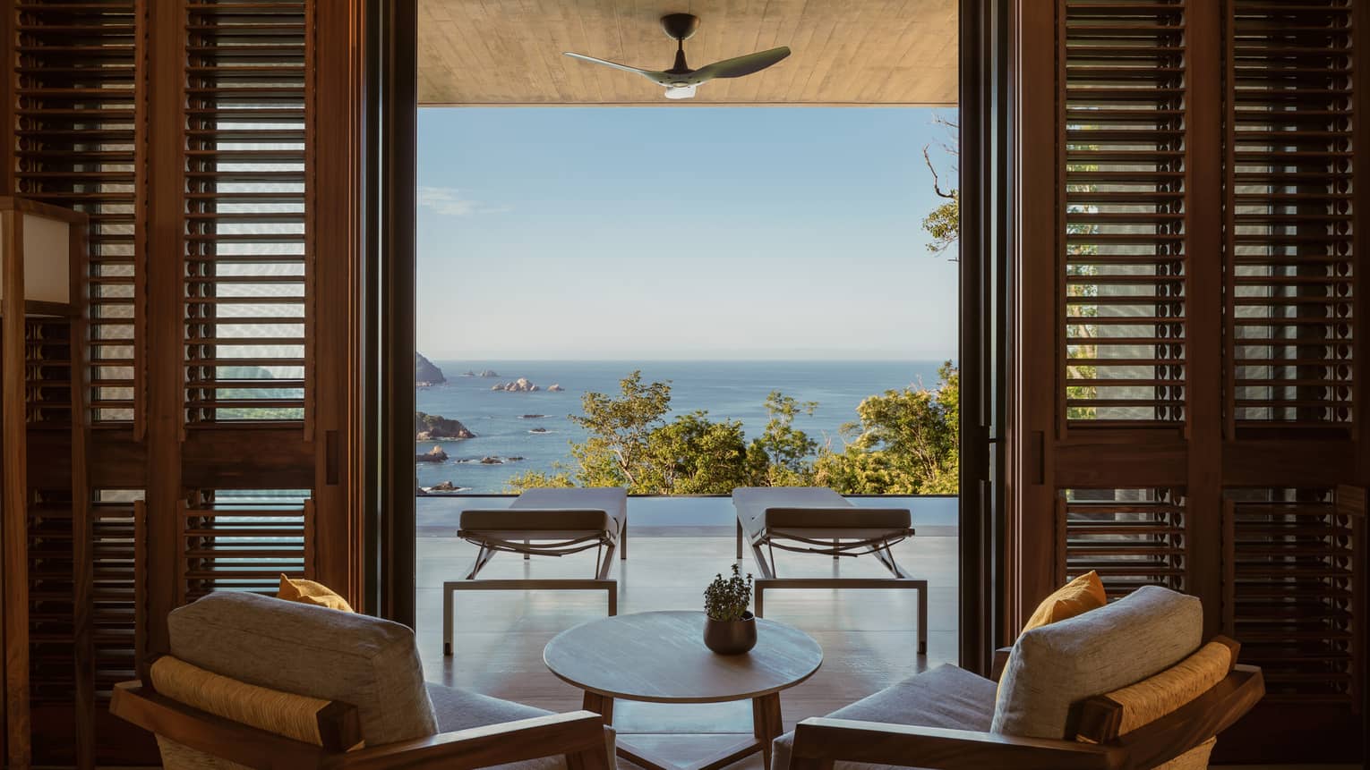 Two arm chairs and small table face outwards to pool terrace, overlooking trees and ocean