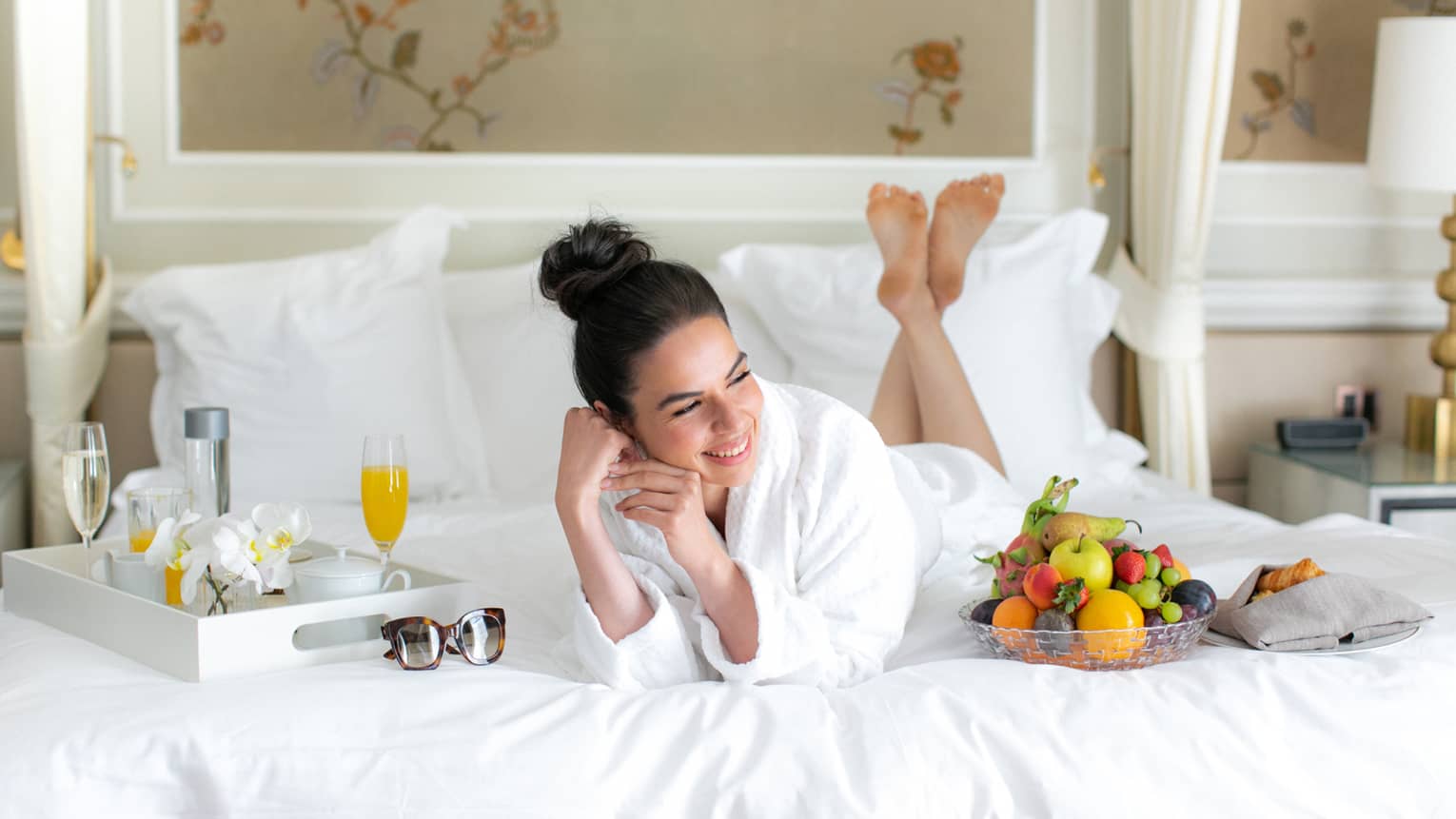 A woman in a spa robe laying on a bed with juice and fruit.